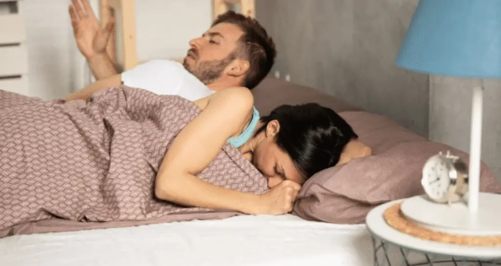 Couple in bed appearing distressed, with woman turned away and man looking frustrated, highlighting emotional impact of painful intimacy.