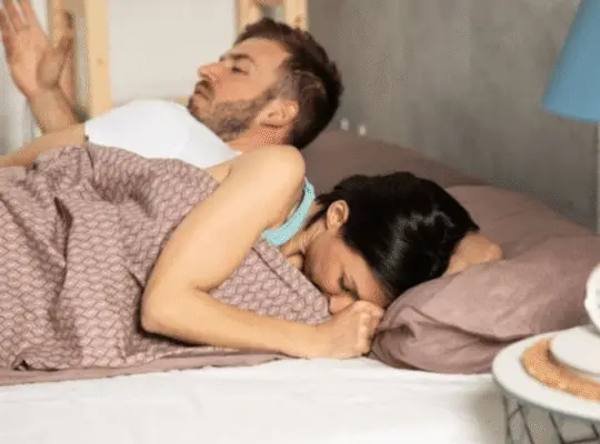 Couple in bed appearing distressed, with woman turned away and man looking frustrated, highlighting emotional impact of painful intimacy.