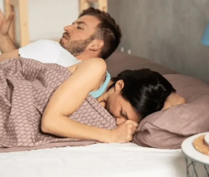 Couple in bed appearing distressed, with woman turned away and man looking frustrated, highlighting emotional impact of painful intimacy.