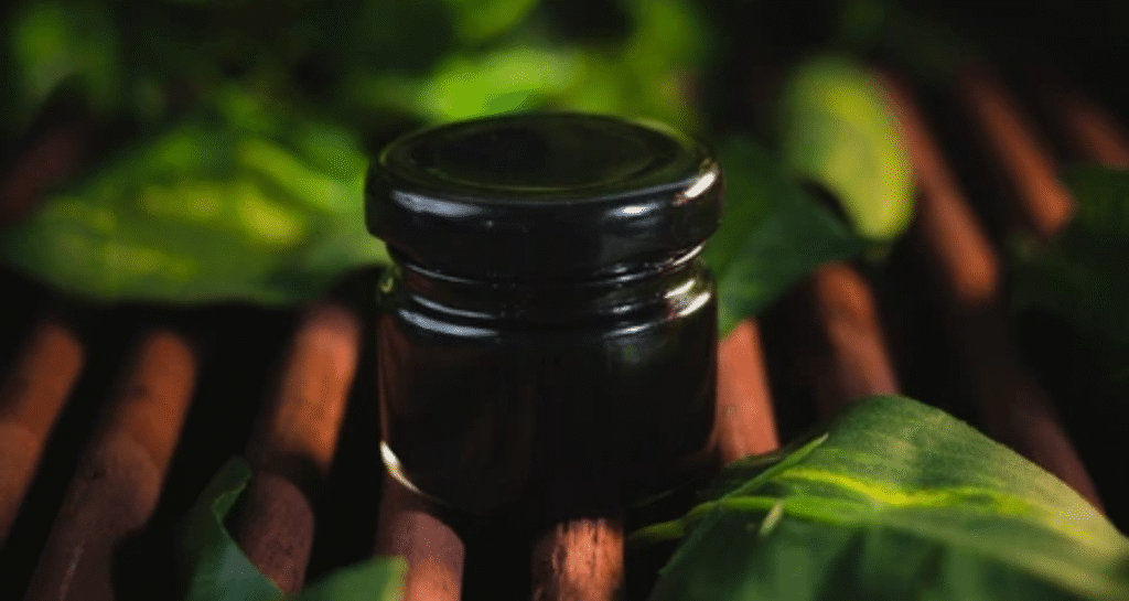 Dark glass jar containing pure shilajit resin surrounded by green leaves on wooden surface
