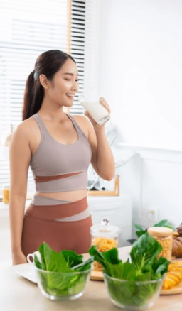 Woman in activewear drinking a glass of milk surrounded by healthy foods on a kitchen table .